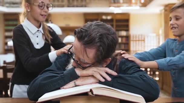 Schöne Teenager Schulkinder wecken ihren müden respektierten Großvater, der auf Büchern in der Bibliothek schläft — Stockvideo