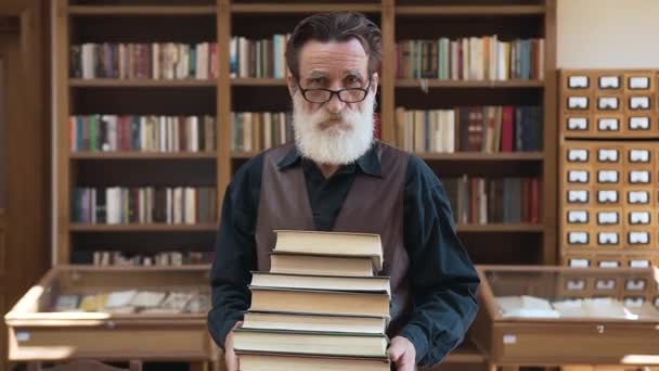 Уважаемый старший бородатый мужчина держит в руках много книг и позирует перед камерой на книжных полках — стоковое видео