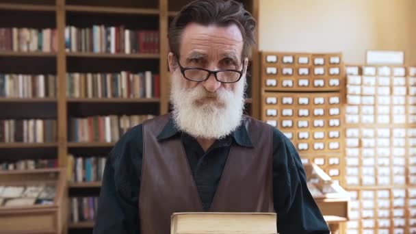 Esteemed old bearded man in glasses holding books in hands and looking at camera on the bookshelves background — Stock Video