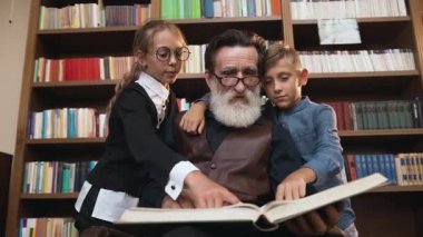 İlgili büyükbaba ve torunlarının aile portresi. Kitaplıkların yanında oturmuş büyüteçle kalın kitaplar okuyorlar.