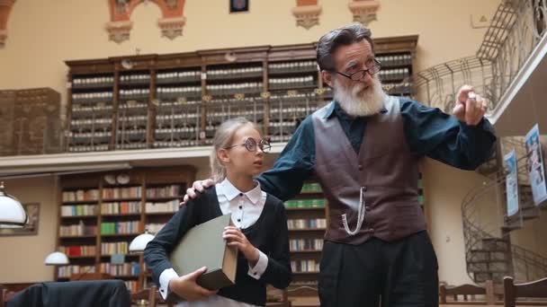 Розумний старший бородатий чоловік розповідає про бібліотеку своїй милій зацікавленій онуці під час прогулянки всередині бібліотеки — стокове відео