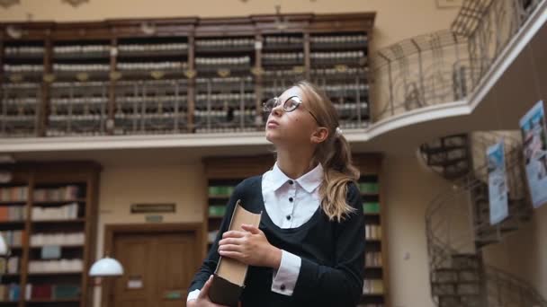 Nahaufnahme des schönen blonden Teenie-Mädchens in einer Brille, die ein dickes Buch hält und durch den Bibliotheksraum kommt — Stockvideo
