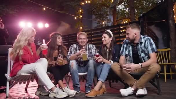 Gran tiempo con amigos. Cuatro jóvenes alegres charlando y bebiendo cerveza mientras están sentados en las sillas en la casa de atrás del patio en la noche de fin de semana — Vídeo de stock