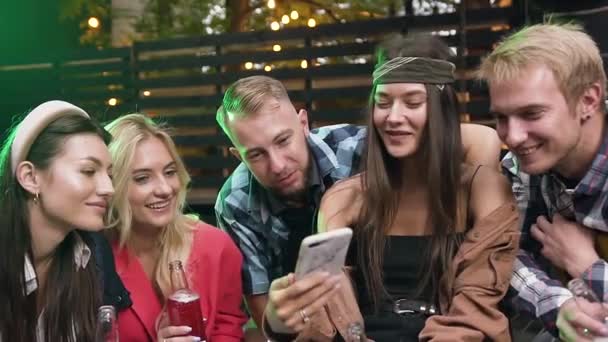 Retrato de una joven hermosa mujer usando la aplicación muestra cosas interesantes en su teléfono inteligente a sus cuatro amigos mientras se divierten en la fiesta el fin de semana. Al aire libre — Vídeos de Stock