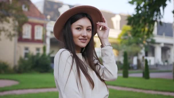 Primer plano de la encantadora feliz dama alegre con el pelo largo y castaño y hermoso sombrero que posando en la cámara con linda sonrisa delante de maravillosas viviendas — Vídeos de Stock