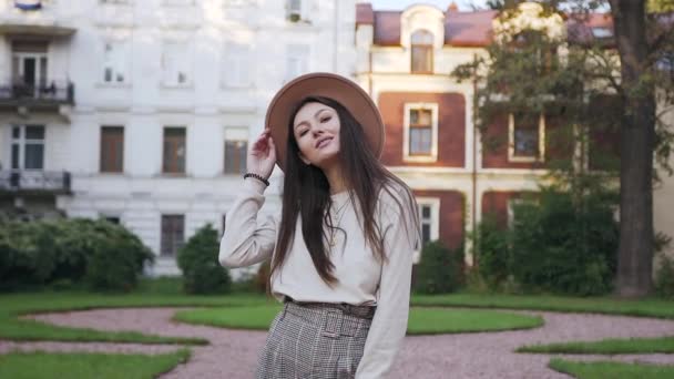 Веселая симпатичная 30-летняя женщина в модной шляпе позирует на камеру с искренней улыбкой перед красивыми особняками — стоковое видео