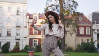 Güzel görünüşlü, güler yüzlü, modern 30 'lu yaşlarda şık kıyafetler ve şapkalı bir kız. Telefonunda poz verip zengin köşklerin arka planında komik fotoğraflar çekiyor.