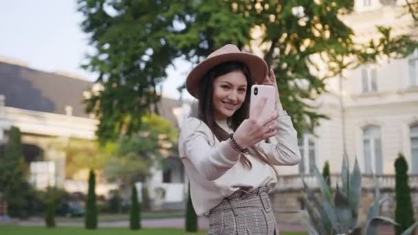 Atractivo retrato de encantadora dama alegre feliz en sombrero de moda y ropa que hace autofoto divertida en su teléfono inteligente durante el paseo cerca de lujosas viviendas — Vídeos de Stock