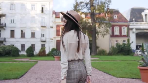 Vue arrière de magnifique femme heureuse et gaie en chemisier et pantalon de mode et chapeau beige qui marche à proximité des demeures luxueuses et posant sur la caméra avec un joli sourire — Video