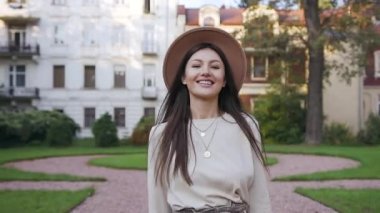 Güzel malikanelerin yanındaki yeşil yolda yürüyen şık elbiseli ve şapkalı mutlu, neşeli genç bayan ve güzel bir gülümsemeyle kameraya bakıyor.