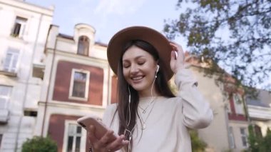 Uzun kahverengi saçları ve moda şapkasıyla evin bahçesindeki yeşil ağacın yanında duran ve kulaklıklarıyla güzel müziğin keyfini çıkaran büyüleyici genç kadın.