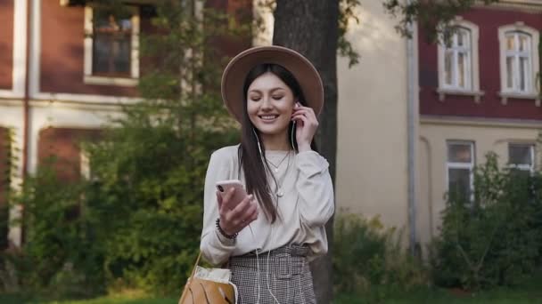 Счастливая веселая красивая женщина в бежевой шляпе слушает хорошую музыку на наушниках во время прогулки возле красивого особняка — стоковое видео
