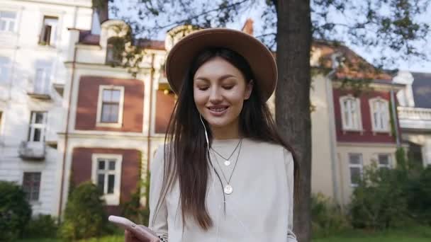 Romantik, çekici, hoş şapkalı genç bir kadın güzel bir evin yanında yürüyor ve kulaklıklarıyla müzik dinliyor. — Stok video