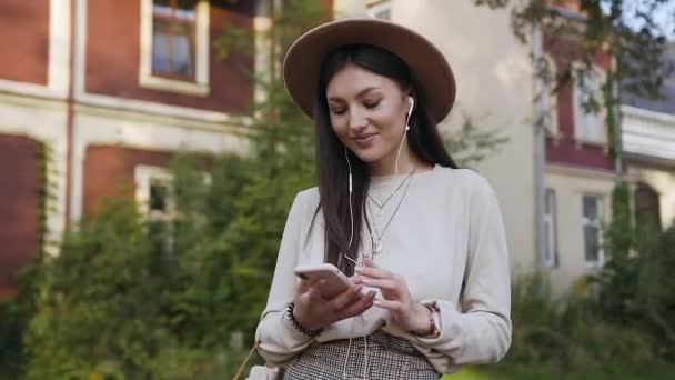 Vista frontal de la hermosa mujer feliz sonriente en sombrero beige caminando cerca de la hermosa mansión y buscando una canción agradable en su lista de reproducción para escuchar en los auriculares — Vídeo de stock