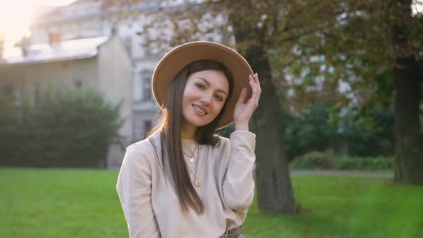 Preciosa mujer feliz sin preocupaciones en sombrero de moda mirando a la cámara con una sonrisa sincera en el fondo de árboles verdes y hermosas casas — Vídeos de Stock