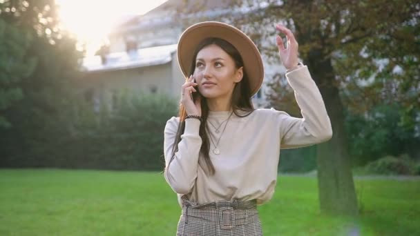 Mosolygó csinos fiatal nő bézs kalapban áll a közelben zöld fa a házak udvarán, és telefonon beszél — Stock videók