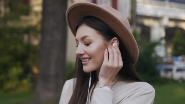 Chica de moda impresionante alegre de 30 años en sombrero elegante con auriculares y disfrutar de la música al estar de pie en el patio de las casas — Vídeo de stock