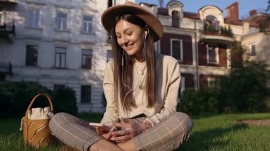 Moda şapkalı, güler yüzlü, neşeli bir bayan evlerin yakınındaki çimenlerde oturuyor ve telefondaki müzik listesini izliyor kulaklıkla dinlemek için.