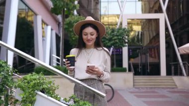 Model şapkalı güzel, neşeli genç bir kadın ofis merkezinin yanında duruyor cam pencereli ve telefon kullanırken kahve içiyor.