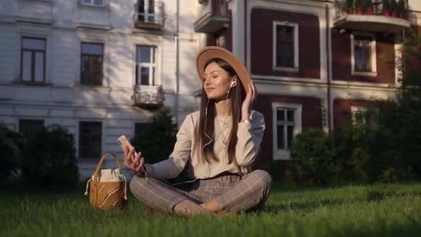 Hermosa joven esbelta mujer satisfecha en sombrero modish sentado sobre hierba verde cerca de casas acogedoras y escuchar música en los auriculares — Vídeos de Stock