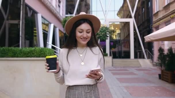 Attraente felice donna soddisfatta in cappello moderno a piedi vicino all'edificio dell'ufficio con caffè di strada e usando il suo telefono mentre beve caffè fresco — Video Stock