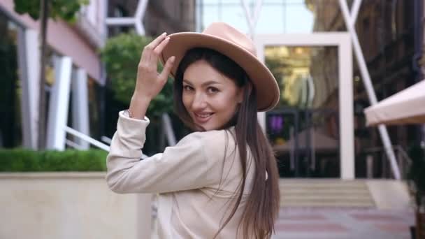 Belle jeune femme heureuse aux longs cheveux bruns vêtus d'une belle blouse et d'un chapeau élégant regardant la caméra pendant sa promenade près d'un bâtiment moderne — Video