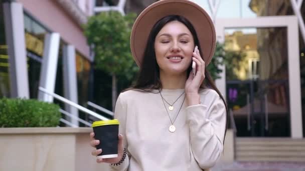 Vista frontale di attraente ragazza euforica in bel cappello con il suo caffè fresco in mano parlando al telefono mentre si avvicina a un bellissimo edificio con pareti di vetro — Video Stock