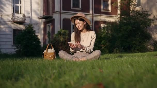 Hermoso retrato de mujer sonriente feliz de buen aspecto con sombrero elegante que se relaja en el césped cerca de las casas y el uso de su teléfono inteligente — Vídeos de Stock