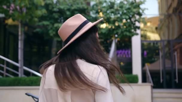 モダンな建物の背景にカメラを置きながら回転しながら、モディッシュな帽子で笑顔の若い女性をアピール — ストック動画
