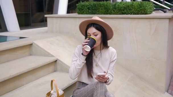 Закрыть очаровательную мечтательную барышню в модной шляпе и стильной одежде, которая наслаждается свежим кофе, сидя на ступеньках возле современного здания — стоковое видео