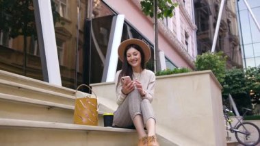 Modern elbiseli, şık şapkalı güzel, narin bir bayan şehir binalarının yakınındaki merdivenlerde oturuyor ve akıllı telefonunu kullanıyor.