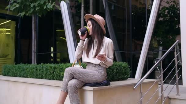 Stílusos modern lenyűgöző nő divatos kalap ül beton mellvéden közelében városi épület, ivás friss kávé és okostelefon használata — Stock videók