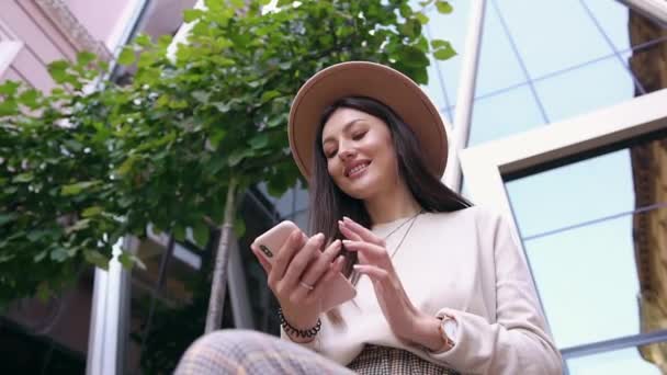 Вниз вид очаровательной веселой женщины в модной шляпе, сидящей возле больших красивых зданий и прокручивающей свой смартфон — стоковое видео
