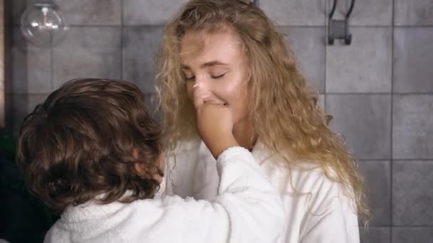 Criança pequena com cabelo encaracolado colocando mancha com creme de cuidados da pele no rosto das mães e, em seguida, mãe abraçando-o com amor — Vídeo de Stock