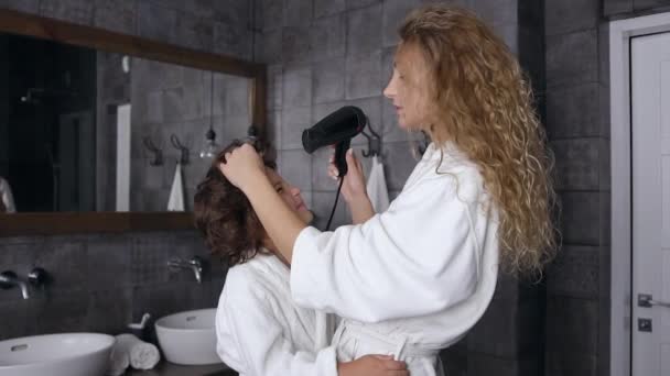 Splendida giovane donna bionda con i capelli ricci in vestaglia bianca asciugando i capelli dei suoi figli in bagno — Video Stock