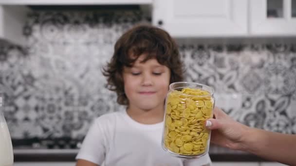 Snygg liten pojke som sitter vid bordet och väntar medan hans mamma fyller skålen med majsflingor — Stockvideo