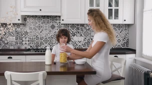 Belo retrato de mãe encantadora e filho sorridente que sentado na mesa da cozinha e criança assistindo como a mãe enchendo seu prato com flocos de milho — Vídeo de Stock
