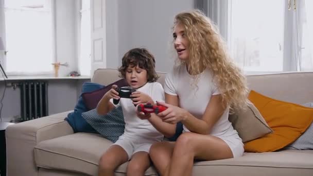 Pretty cheerful blond woman with curly hair playing video game with her attractive little son using joysticks,sitting on the cozy couch in the room — ストック動画