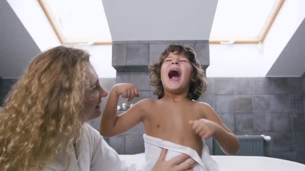 Usmívající se pohledný chlapec ukazuje paže gesta, jak je silný, zatímco jeho roztomilé blond matka otírá tělo ručníkem — Stock video