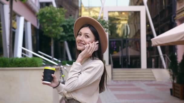 Ganska glad 30-årig kvinna i modisk hatt vänder sig när du går nära vackra moderna byggnad och talar i telefon med kvinnliga vän — Stockvideo