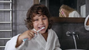 Siyah kıvırcık saçlı gülümseyen küçük çocuk banyodaki aynanın yanında dişlerini fırçalıyor.