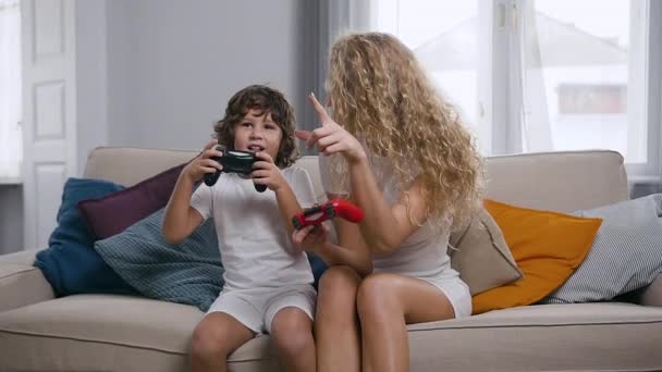 Симпатичная счастливая мама и активный милый мальчик, сидящий на мягком диване и пользующийся джойстиками во время игры — стоковое видео