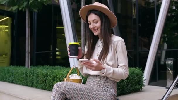 Повільний рух чарівної романтичної молодої леді в стильному капелюсі і стильному одязі, який сидить на бетонному парапеті, використовуючи свій телефон і п'є каву — стокове відео