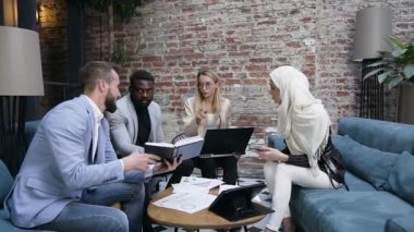 Ofis kanepelerinde oturup kitap, bilgisayar ve raporlar kullanarak ortak projeyi tartışan çok ırklı ciddi iş adamlarının çekici portresi.