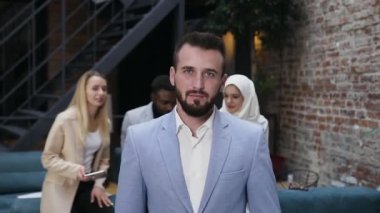 Yakışıklı, sakallı, mavi ceketli genç bir iş adamı. Ofisteki iş arkadaşlarının arka planlarına bakıyor.