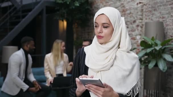 Atraente mulher de negócios muçulmana de 35 anos em hijab digitando no i-pad e de pé no escritório moderno no fundo de seus parceiros de negócios multirraciais — Vídeo de Stock