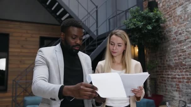 Schöner bärtiger dunkelhäutiger Geschäftsmann, der mit seiner blonden, atemberaubenden Kollegin verschiedene Dokumente durchsieht, während er durch das Büro geht — Stockvideo