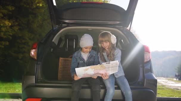 Γοητευτικό ξανθό κοριτσάκι με αστεία ακουστικά που δείχνει το δρόμο για τον αδελφό της όπου πηγαίνουν με τους γονείς το Σαββατοκύριακο, ενώ κάθονται στο πορτ-μπαγκάζ των αυτοκινήτων — Αρχείο Βίντεο