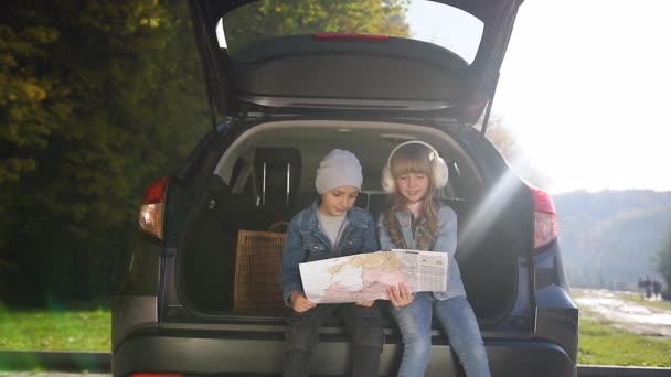 Gyönyörű örömteli gyerekek ülnek a csomagtartóban turista térképpel és találgatással, hogy melyik úton mennek hétvégére a szüleikkel. — Stock videók