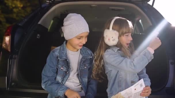 Привлекательный мальчик и девочка сидят в багажнике машины и спорят, в каком направлении ехать на выходные с родителями — стоковое видео